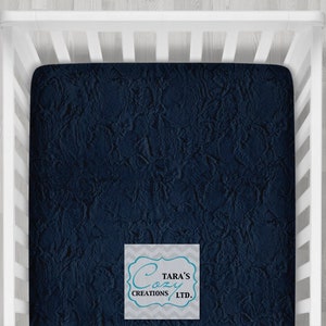 May include: A navy blue fitted cot sheet with a textured, fuzzy surface. The sheet is made by Tara's Cozy Creations LTD.