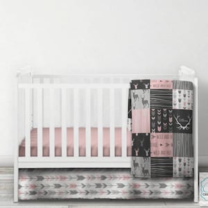 May include: A white cot with a pink and grey patterned cot skirt and a pink and grey patterned blanket. The blanket has the text "Free Wild and Free" on it.