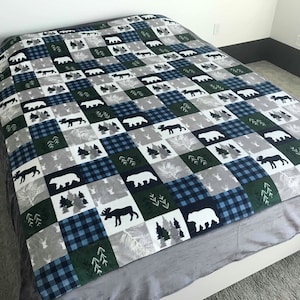 May include: A soft, plush blanket with a patchwork design featuring bears, moose, and trees in shades of blue, green, and white. The blanket is laid out on a bed with a gray headboard.