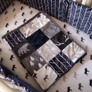 May include: A baby crib set with a patchwork quilt featuring bear, antler, and arrow designs in grey, white, and navy blue. The crib sheets have a moose pattern, and the bumper pads have a chevron pattern.