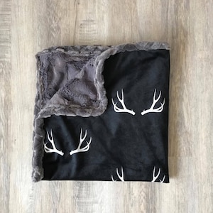 May include: A black and gray fleece blanket with a white deer antler pattern. The blanket is soft and cozy, perfect for snuggling up on the couch.
