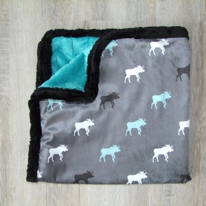 May include: A gray and teal reversible baby blanket with a black faux fur trim. One side features a pattern of white moose silhouettes on a gray background. The other side is teal with a geometric pattern.