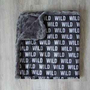 May include: A black and gray baby blanket with a soft, plush texture. The blanket features a repeating pattern of the word "WILD" in white.