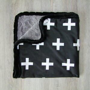 May include: Black and white baby blanket with a plush, soft texture. One side features a white cross pattern on a black background. The other side has a gray arrow pattern.
