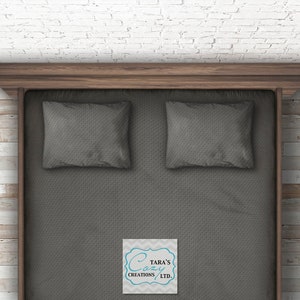May include: A grey bed with two grey pillows on top. The bed is made with a grey textured fabric. The bed is in a room with white brick walls and wood floors. The text "Tara's Cozy Creations LTD." is on a white tag in the bottom right corner of the image.