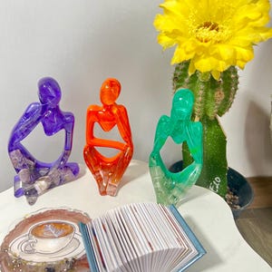May include: A collection of decorative resin art pieces. Includes three colorful human figure sculptures in purple, orange, and green. Also shown are a coaster with a coffee design and a small blue book with white pages. A cactus with a large yellow flower is in the background.