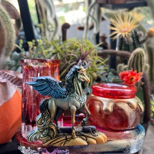 May include: A detailed figurine of a winged unicorn with blue wings and a flowing mane stands on a decorative base. A red and gold jar and a pink glass vase are also visible, surrounded by plants and cacti.