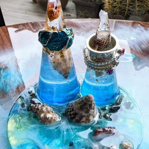 May include: A decorative resin art piece with two lighthouse-shaped structures. The structures are made with blue resin and embedded seashells. The base is a round, ocean-themed design with seashells, a starfish, and blue and white resin. The lighthouses have a blue and gold trim.