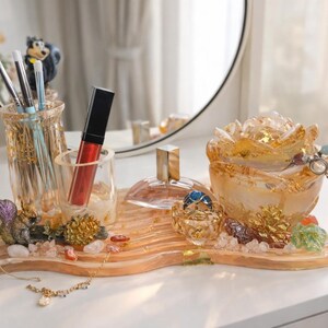 May include: A decorative vanity tray with a clear glass perfume bottle, a red lipstick, and a rose-shaped jewelry holder. The tray also holds makeup brushes and a small figurine. A necklace and rings are also visible.