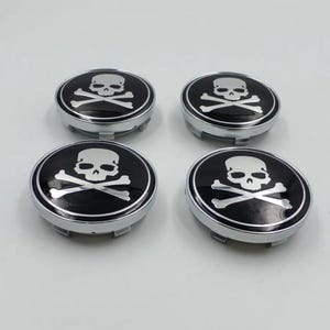 May include: Four black and silver skull and crossbones hubcaps. Each hubcap features a white skull and crossbones design on a black background, surrounded by a silver rim. The hubcaps are circular and have a metallic finish.