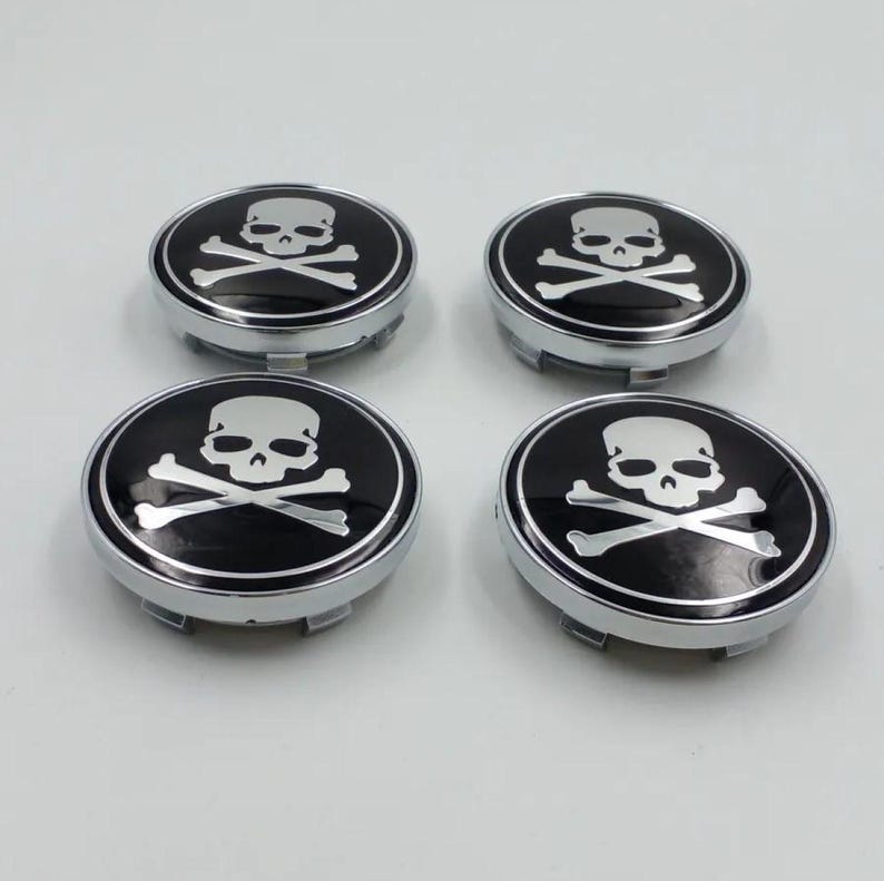 May include: Four black and silver skull and crossbones hubcaps. Each hubcap features a white skull and crossbones design on a black background, surrounded by a silver rim. The hubcaps are circular and have a metallic finish.