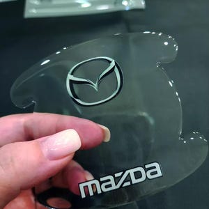 Könnte beinhalten: Transparenter, gebogener Türgriffschutz für Autos mit dem Mazda-Logo in Schwarz und Weiß. Das Wort "Mazda" ist weiß gedruckt. Der Schutz wird von einer Hand gehalten.