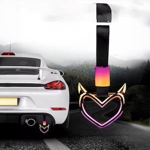 Peut inclure: Accessoire de voiture en forme de cœur avec des cornes de diable. Le cœur présente un dégradé de rose, de violet et d'or. Il est attaché à une sangle noire. Une voiture de sport blanche est visible en arrière-plan.