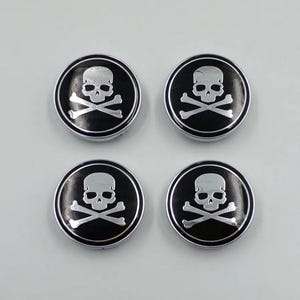 May include: Four circular, black and silver skull and crossbones emblems. Each emblem features a silver skull and crossbones design set against a black background, surrounded by a silver border. The emblems are arranged in a square.