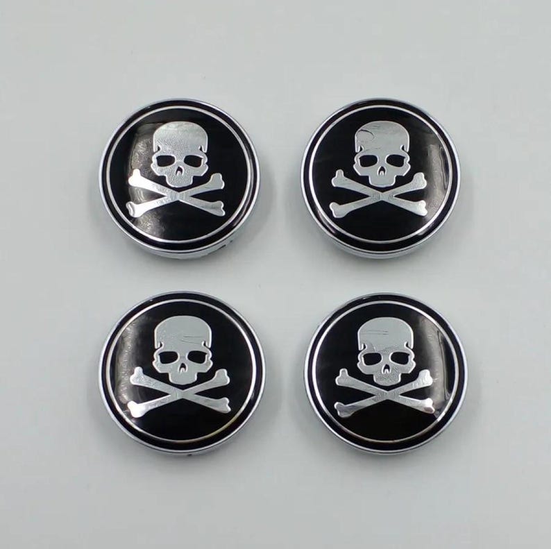 May include: Four circular, black and silver skull and crossbones emblems. Each emblem features a silver skull and crossbones design set against a black background, surrounded by a silver border. The emblems are arranged in a square.