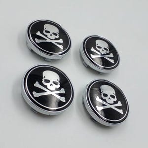 May include: Four black and silver skull and crossbones wheel center caps. Each cap features a white skull and crossbones design on a black background, surrounded by a silver border. The caps are round and have a metallic finish.