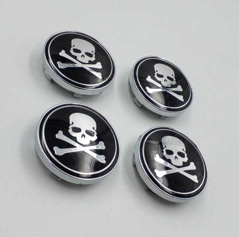 May include: Four black and silver skull and crossbones wheel center caps. Each cap features a white skull and crossbones design on a black background, surrounded by a silver border. The caps are round and have a metallic finish.