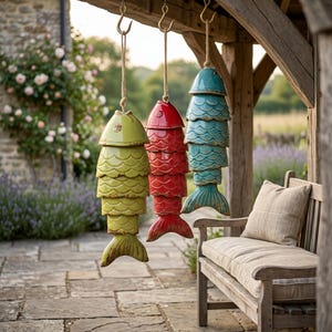 Peut inclure: Trois carillons en forme de poissons en céramique, verts, rouges et turquoise, suspendus à une structure en bois. Chaque poisson est fait d'écailles superposées et possède une nageoire caudale. Les carillons sont suspendus par des cordes et des crochets en métal.