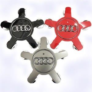May include: Three car wheel centre caps in black, red, and silver. Each cap features a five-point star design with a silver Audi logo in the centre. The caps have a glossy finish and are designed for automotive use.