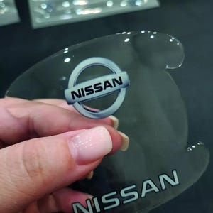May include: Clear, curved Nissan logo emblem. The emblem features the Nissan logo in a silver and blue circle, with the word "NISSAN" printed in silver. The emblem is held in a hand.