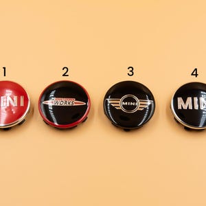 Peut inclure: Quatre enjoliveurs de roue MINI noirs et rouges avec différents logos. Les enjoliveurs sont ronds et ont une finition brillante. Le premier enjoliveur est rouge avec "MINI" en argent. Le deuxième enjoliveur est noir avec "JOHN COOPER WORKS" en blanc et rouge. Le troisième enjoliveur est noir avec un logo MINI argenté. Le quatrième enjoliveur est noir avec "MINI" en argent.