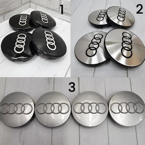 Może przedstawiać: Kolekcja czarnych i srebrnych kołpaków Audi. Kołpaki posiadają logo Audi, zestaw czterech zazębiających się pierścieni, w kontrastowym kolorze. Kołpaki są okrągłe i wydają się być wykonane z tworzywa sztucznego lub metalu.