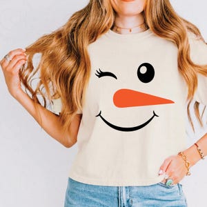 May include: Cream-colored t-shirt featuring a snowman face design. The design includes a winking eye, a black eye, an orange carrot nose, and a smiling mouth. The shirt is paired with blue jeans.