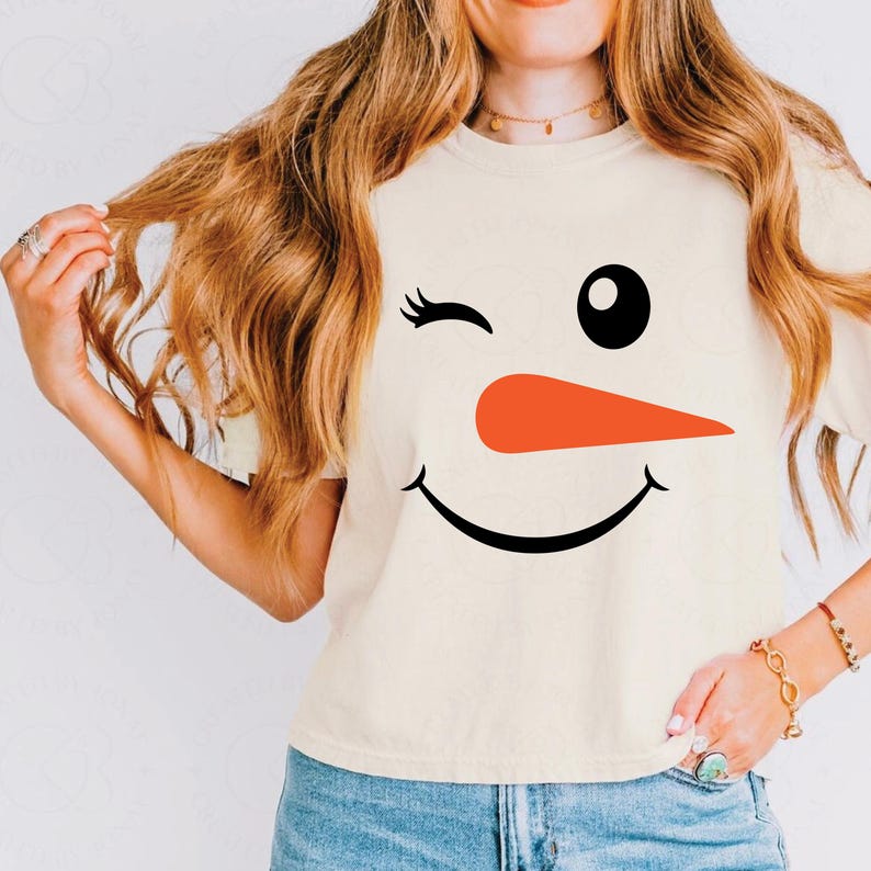 May include: Cream-colored t-shirt featuring a snowman face design. The design includes a winking eye, a black eye, an orange carrot nose, and a smiling mouth. The shirt is paired with blue jeans.