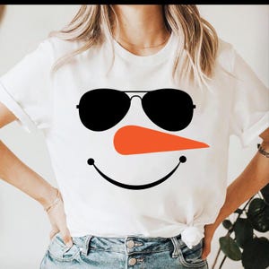 May include: White t-shirt featuring a snowman face design. The design includes black sunglasses, an orange carrot nose, and a black smiling mouth. The shirt is a short-sleeved crew neck.