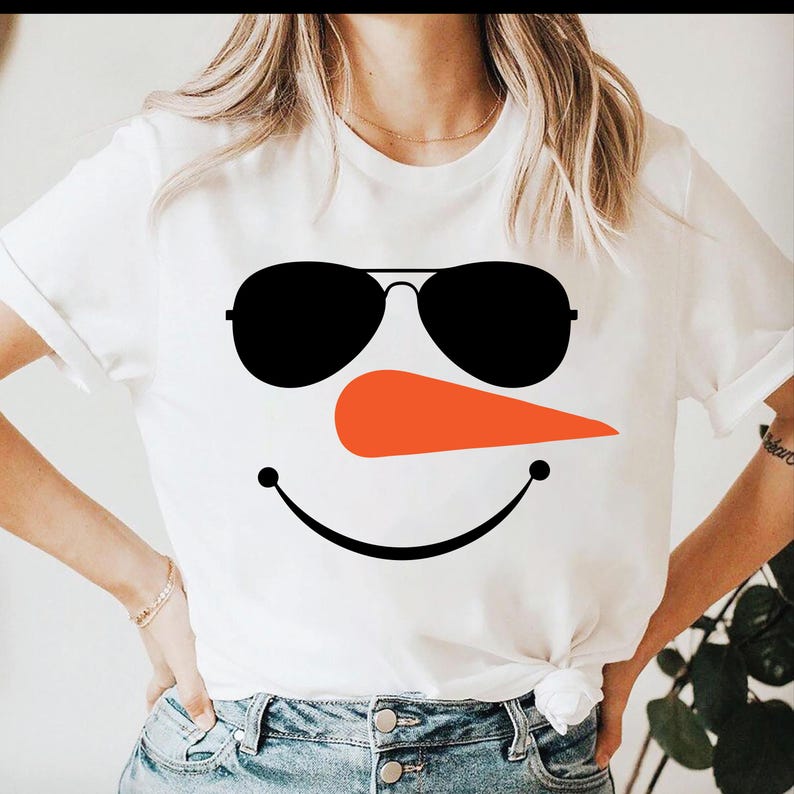 May include: White t-shirt featuring a snowman face design. The design includes black sunglasses, an orange carrot nose, and a black smiling mouth. The shirt is a short-sleeved crew neck.