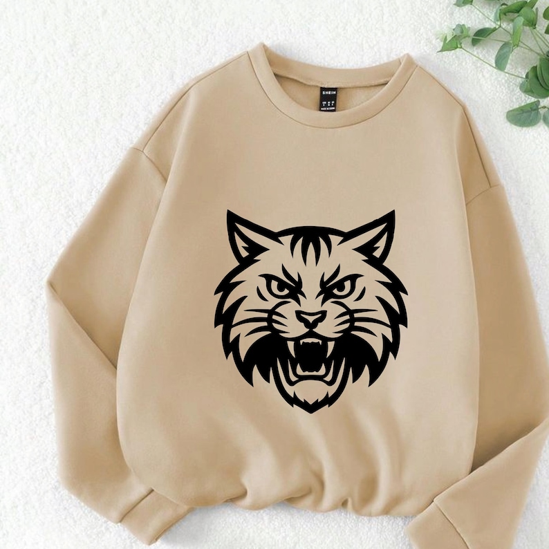 May include: A tan sweatshirt featuring a bold black graphic of a snarling wildcat. The sweatshirt has a crew neck and long sleeves. The wildcat graphic is centered on the front of the sweatshirt.