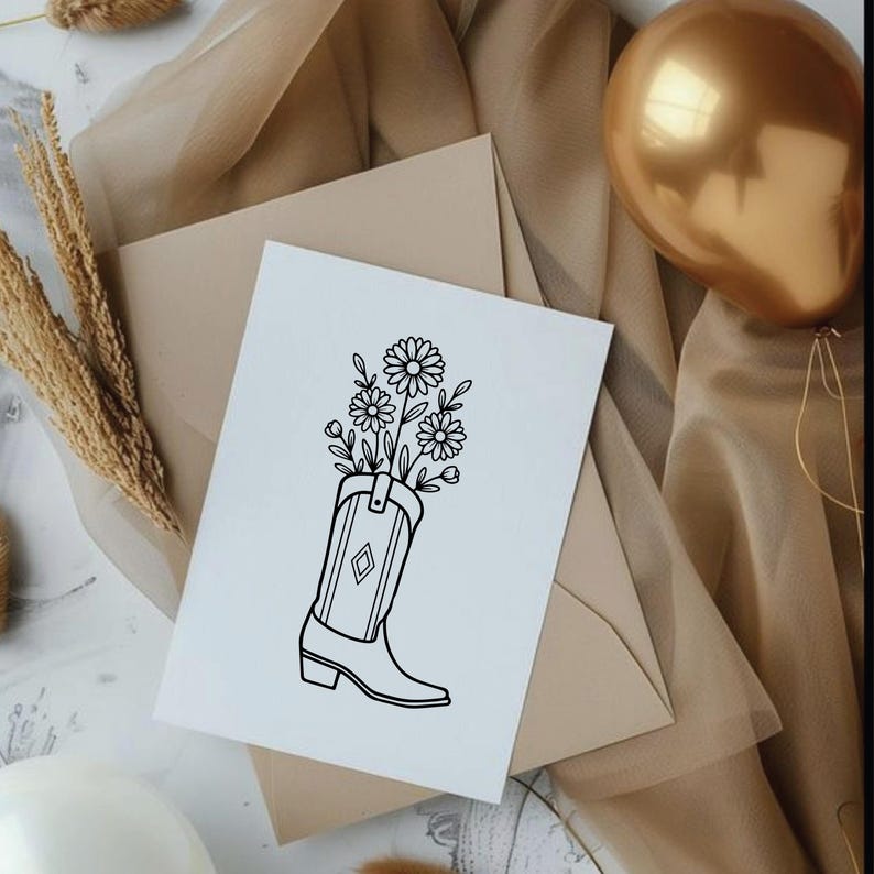 May include: A white card with a black line drawing of a cowboy boot filled with daisies. The card is on top of a tan envelope and a gold balloon is in the background. The boot has a diamond design.