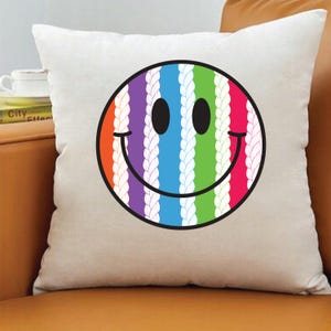 May include: A white square pillow featuring a colorful smiley face design. The smiley face has a black outline and a rainbow-striped interior with a braided rope pattern. The pillow is on a brown surface.