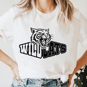 May include: White t-shirt featuring a black and white graphic of a roaring tiger above the word "WILDCATS". The design is centered on the front of the shirt. The shirt is made of a soft, light-colored fabric.