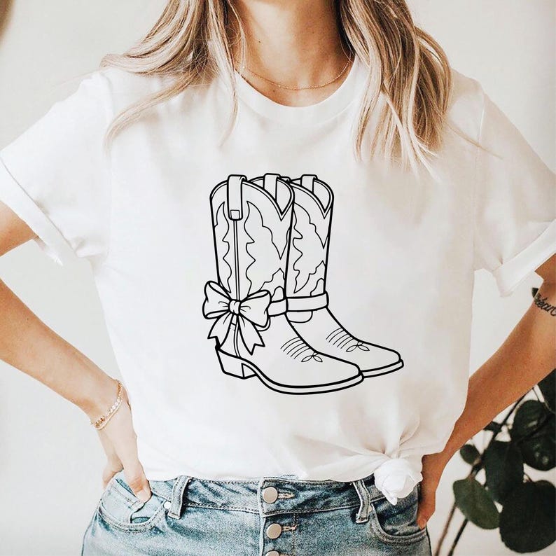 May include: White t-shirt featuring a black and white illustration of a pair of cowboy boots with a bow. The boots have decorative stitching and a bow on the front. The shirt is a casual style.
