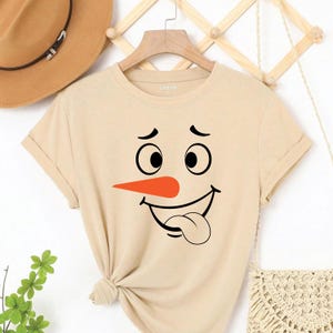 May include: Beige t-shirt featuring a snowman face graphic. The design includes black eyes, a carrot nose, and a smiling mouth with a tongue sticking out. The shirt has rolled-up sleeves and a knot at the bottom.