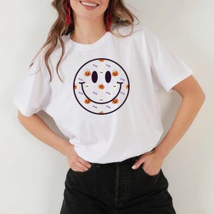 May include: White t-shirt featuring a large smiley face design with a Halloween theme. The smiley face is outlined in black and filled with pumpkins, ghosts, and candy buckets. The shirt has short sleeves and a crew neck.