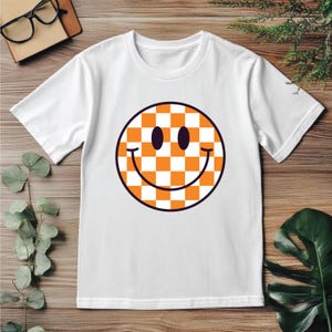 May include: White t-shirt featuring a large, orange and white checkered smiley face with a black outline. The smiley face has two black oval eyes and a curved smile. The shirt is made of a soft material.