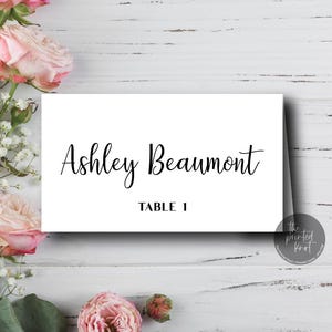 May include: A white place card with the name "Ashley Beaumont" in elegant script and "TABLE 1" in a smaller font. The card is set on a white wooden surface, adorned with pink roses and greenery.