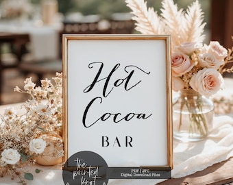 May include: A framed sign with the words "Hot Cocoa Bar" in elegant black script. The sign is in a gold frame and is surrounded by floral arrangements, including roses and pampas grass. The sign is on a table with a white tablecloth.