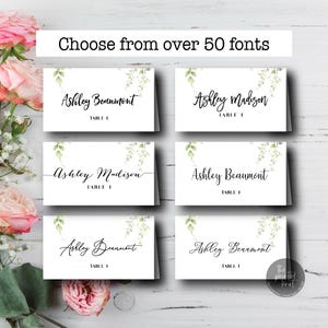 May include: White place cards with elegant calligraphy names and table numbers, accented with watercolor greenery. The cards are displayed on a white wooden surface, with pink roses and greenery in the background. Text reads "Choose from over 50 fonts."