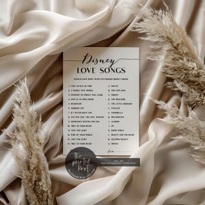 May include: A white card with the text "Disney LOVE SONGS" and a matching game. The card lists Disney songs and movie titles. The card is on a beige fabric background with dried plants.