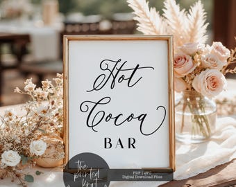 May include: A framed sign with the words "Hot Cocoa Bar" in elegant black script. The sign is in a gold frame and is surrounded by floral arrangements, including roses and pampas grass. The sign is on a table with a white tablecloth.