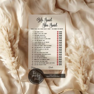 May include: A white "He Said She Said" game card with a list of statements for a bridal shower. The card has a circular logo at the bottom with the text "the printed knot". The game card is on a cream-colored fabric background.