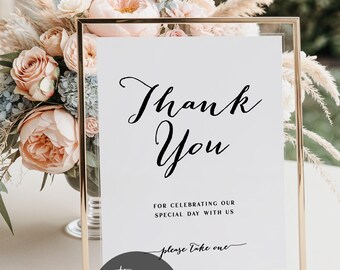 May include: A gold-framed sign with the words "Thank You" in elegant script. The sign includes the text "For celebrating our special day with us" and "please take one." A floral arrangement of pink and cream flowers is in the background.