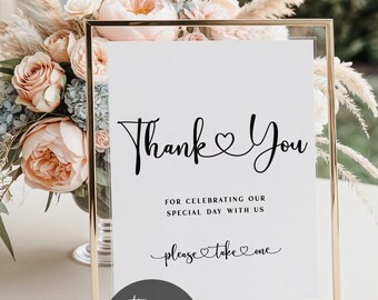 May include: A framed white sign with gold trim, featuring the words "Thank You" in elegant black script. Below, it reads "For celebrating our special day with us" and "please take one." A floral arrangement is in the background.