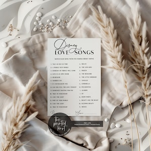 May include: A white card game titled "Disney Love Songs" with a matching game. The card lists Disney songs and movie titles. The card is surrounded by dried plants and sits on a white, textured surface.