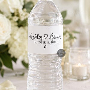 May include: A clear plastic water bottle with a white label featuring the black text Ashley and Brian, the date October 16, 2027, and two heart icons. The label text indicates printed 8.5x2 inch water bottle labels or personalized digital files.