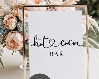 May include: A gold-framed sign with the words "hot cocoa BAR" in black script. The sign is set against a backdrop of soft pink and cream-colored flowers and pampas grass. A circular logo at the bottom reads "The Printed Knot."
