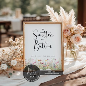 Op de afbeelding: Een ingelijst bord met de tekst "Be Smitten Not Bitten" in elegant schrift, met "Don't Forget The Bug Spray" eronder. De onderkant van het bord heeft een aquarel bloemenontwerp. Het bord staat in een gouden lijst.